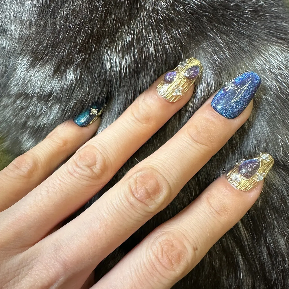 A close-up of a hand with intricately designed nails resting on a textured, gray fur surface. The nails feature a mix of dark blue glitter and gold accents, with small gemstones and teardrop-shaped embellishments, reflecting a celestial and luxurious design.
