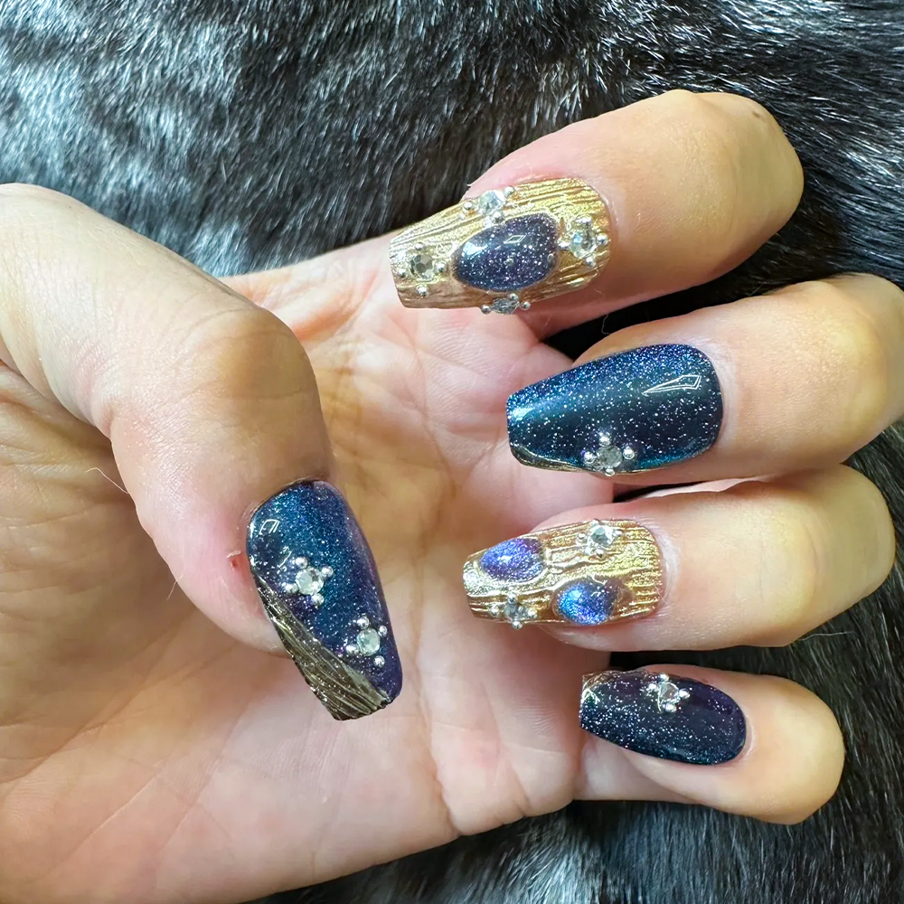 A close-up of a hand with intricately designed nails resting on a textured, gray fur surface. The nails feature a mix of dark blue glitter and gold accents, with small gemstones and teardrop-shaped embellishments, reflecting a celestial and luxurious design.